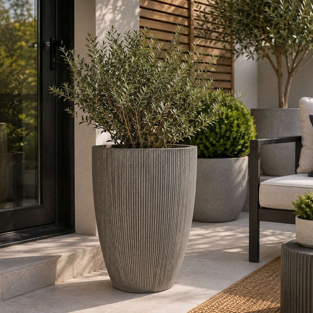 A tall ribbed gray concrete planter holding a leafy green shrub, placed on a patio beside a black-framed glass door, with other outdoor planters and seating in the background.