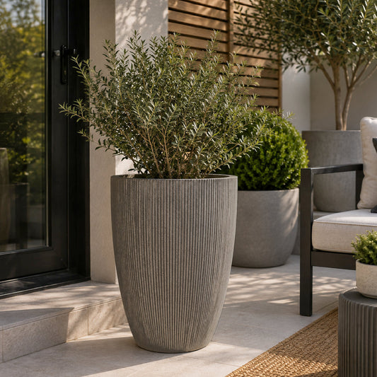 A tall ribbed gray concrete planter holding a leafy green shrub, placed on a patio beside a black-framed glass door, with other outdoor planters and seating in the background.