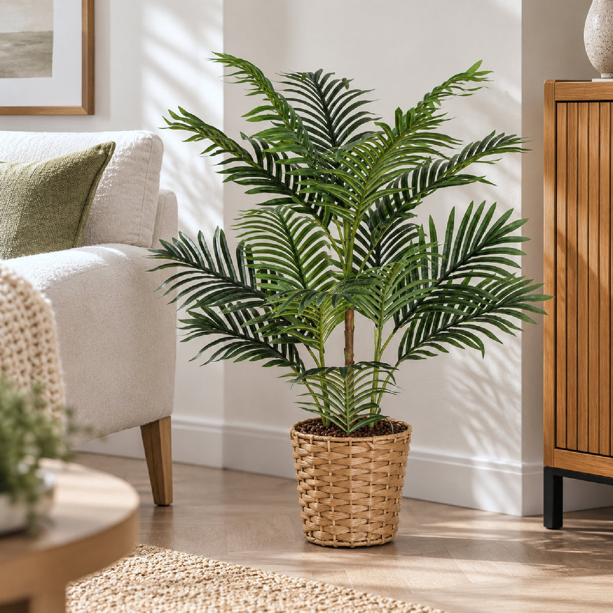 A tall artificial palm-like houseplant with dark green fronds stands in a woven wicker basket pot on a light wood floor in a bright living room setting.