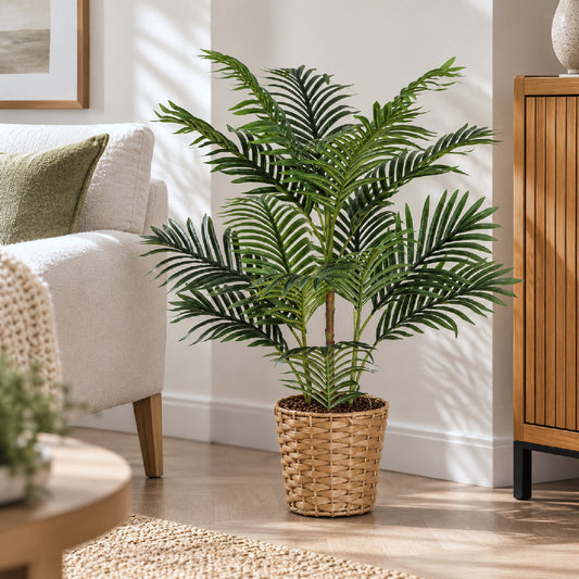 A tall artificial palm-like houseplant with dark green fronds stands in a woven wicker basket pot on a light wood floor in a bright living room setting.