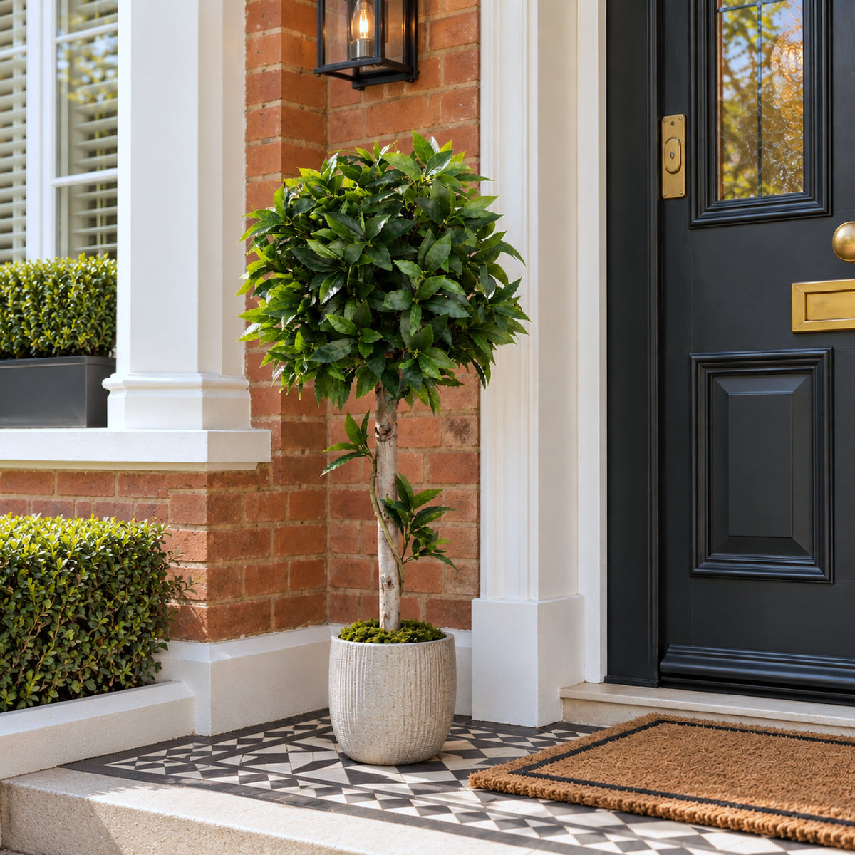 A potted artificial boxwood topiary tree with lush green leaves stands on a patterned stone doorstep next to a black front door and brick wall.