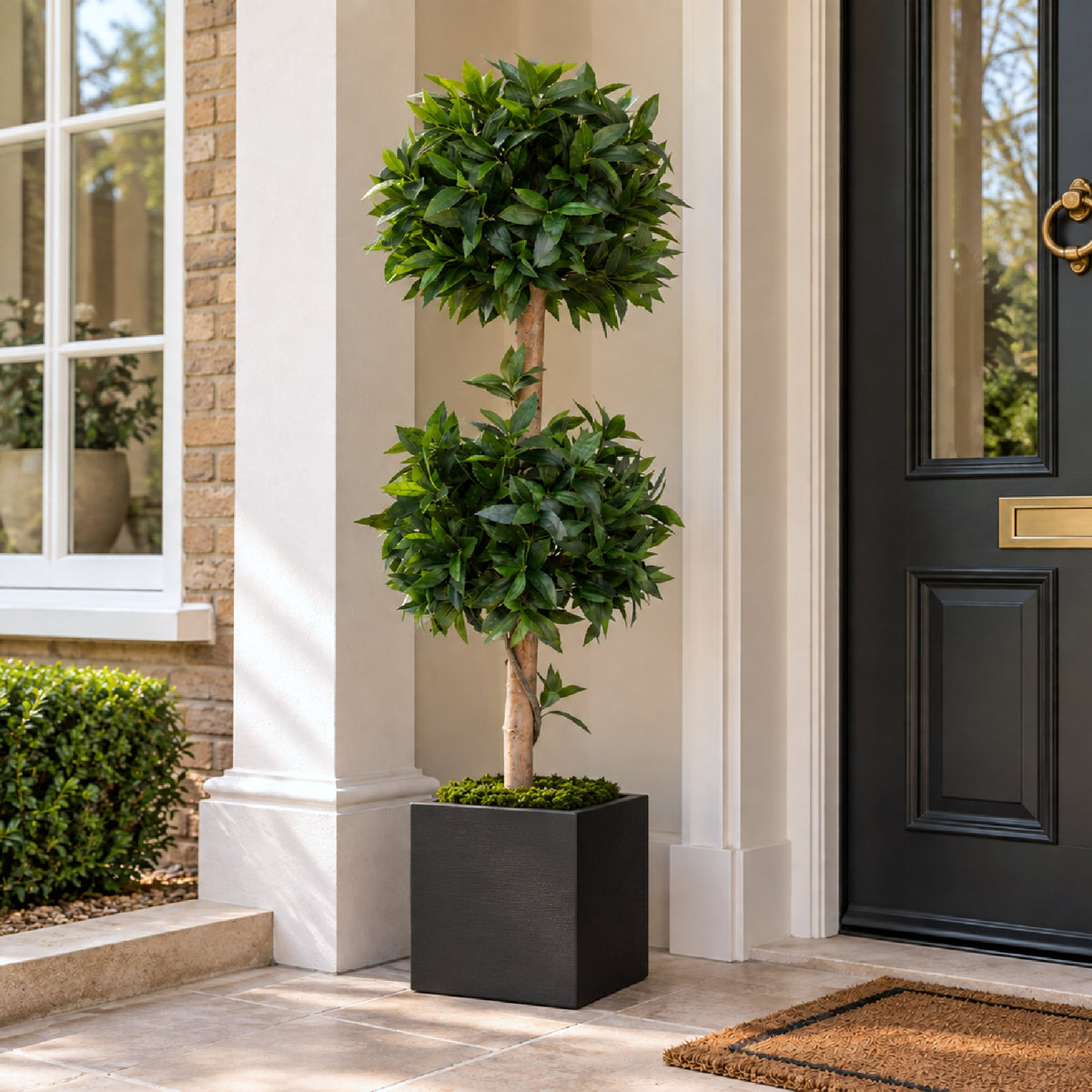 A tall artificial topiary tree with two rounded green plantings on a light wood trunk, displayed in a square black planter on a stone doorstep next to a black front door.