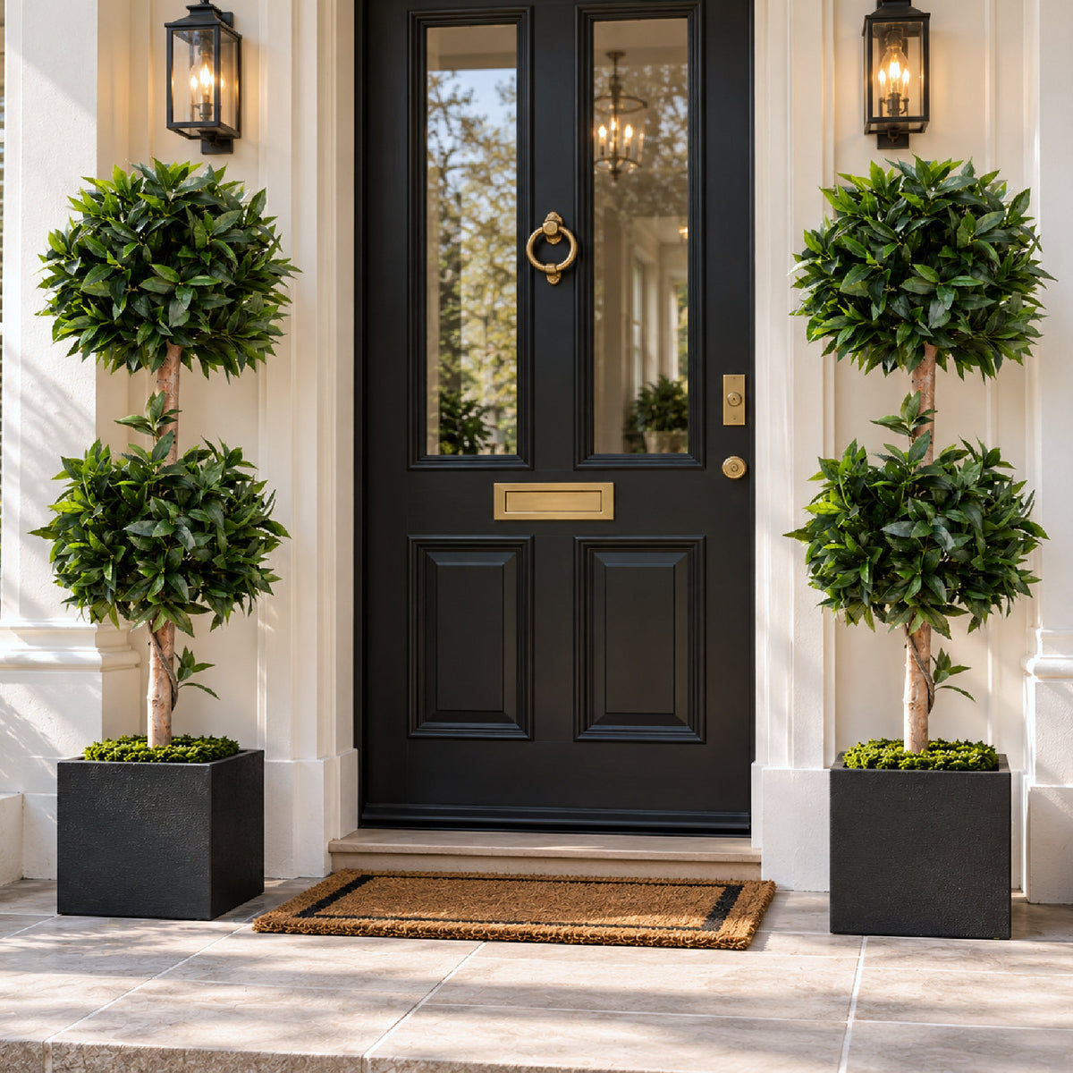 A black front door with gold hardware, flanked by two tall green topiary trees in black square planters on a stone porch.